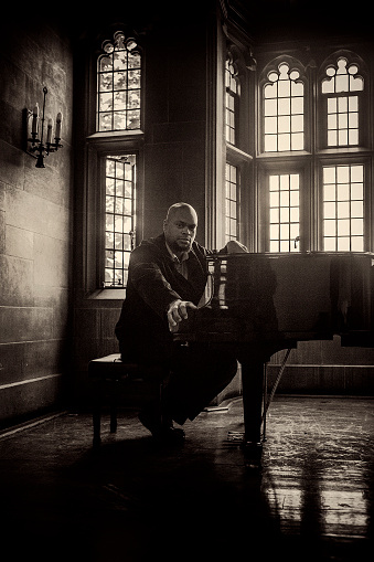 african american man sitting by a classic piano looking at camera, old room with wooden floor and gothic windows