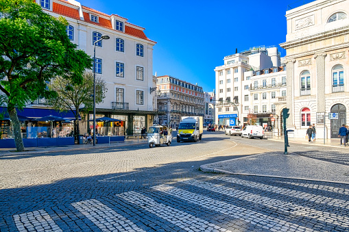 Lisbon, Portugal - January 5, 2023: A bustling street with vehicles driving down it and beautiful story buildings on either side. Pedestrians can be seen on both sides of the sidewalk, and two trees are visible on the left side, with a sunny blue sky in the background.
