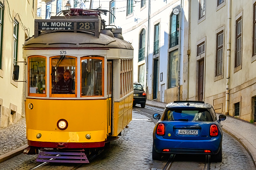 Lisbon, Portugal - January 5, 2023: A tramway surrounded by tall buildings in the daytime, with a yellow tram bus carrying passengers traveling along it. Two cars can also be seen moving in the opposite direction.