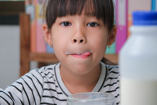 Cute Asian girl drinking a glass of milk at home in living room. Little girl drinking milk in the morning before going to school. Healthy food in childhood.