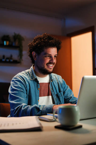 istock Vertical. Happy freelancer man working at home with laptop on a desk at night. Young guy smiling and browsing on internet using a computer at living room. Entrepreneur looking bank finances invests 1484808561