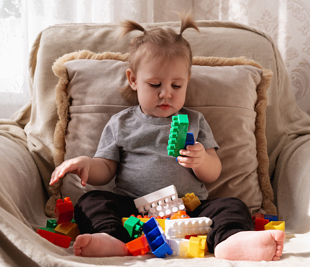 A little sweet sad girl is sitting in an armchair and looking at the toy blocks of the constructor with a bored look.