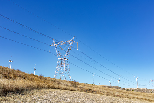 Castilla y León, Spain. High voltage pylon in rural landscape surrounded by wind power pylons against blue sky