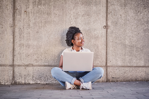 Picture of young woman sitting with laptop against concrete wall in urban environment