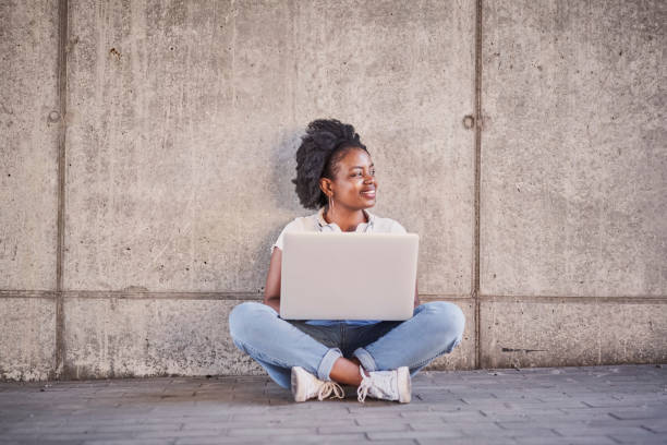 Picture of young woman sitting with laptop against concrete wall in urban environment stock photo