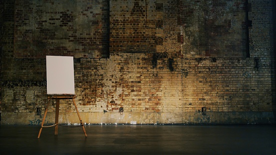 A blank canvas sitting on an easel in front of a rustic brick wall on a theatre stage, with room for text and graphics around the artwork.