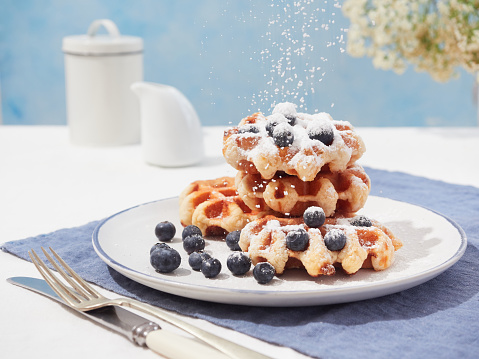 powdered sugar falling sprinkled on top of Belgian waffles with blueberries served on a plate on blue napkin on the table, blue background with copy space.