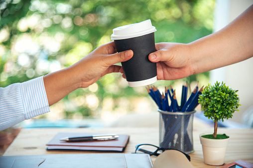 Close up hands business woman colleague drink coffee at office desk together. Friendship teamwork hold caffeine hot tea cup freshness lifestyle. Businesspeople give coffee cup to team active drinking