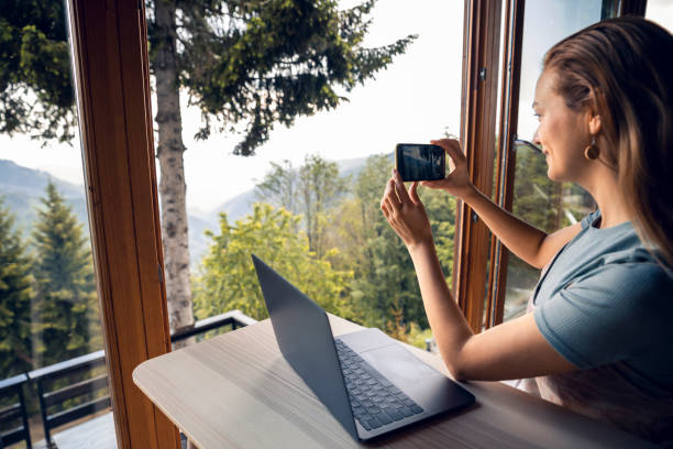 istock Young woman taking a picture through the window of her log cabin while using a laptop 1484847728