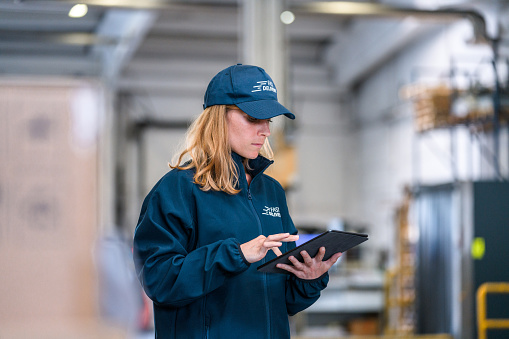 Caucasian female delivery worker using a digital tablet for parcel searching. Wearing blue uniform and standing inside of an airport warehouse.