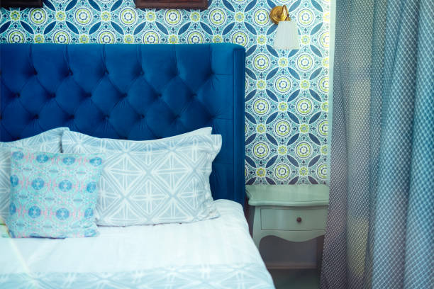 istock Empty bed with light colored linens, pillows with blue patterns and duvet. Classic wall lamp and wallpaper in bright blue geometric particles. 1484852910