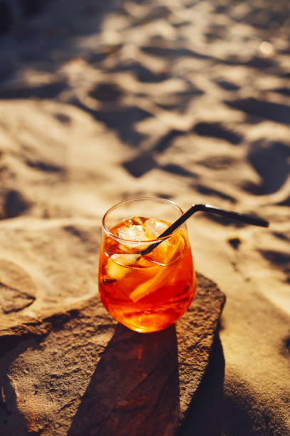 rodaja de naranja y cóctel de aperol spritz sobre una piedra con el telón de fondo de una playa de arena. vaso con agua mineral con gas cítrica helada y paja. - summer party drink umbrella concepts fotografías e imágenes de stock