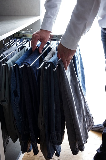 A young adult male standing in an open closet, selecting a pair of jeans to wear.
