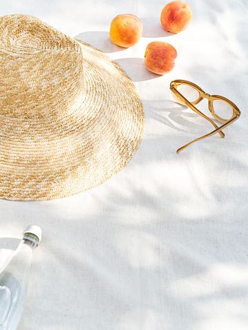 Beach vibe and still life minimalist photo. Tropical summertime picnic in the shadow. A closeup of summer accessories: sunglasses and a straw hat, a bottle of water and peaches refresh the sunny day.