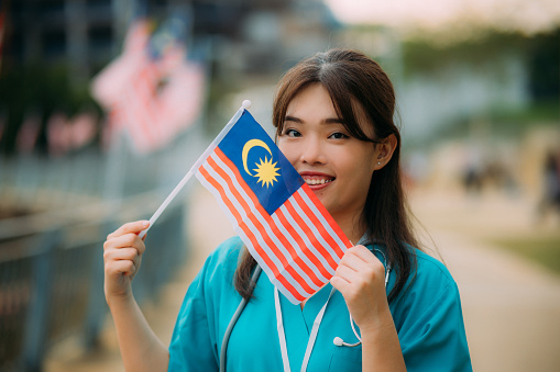 Portrait of a young Asian female medical staff holding the Malaysian flag at a public park, celebrating Independence Day and National Day.