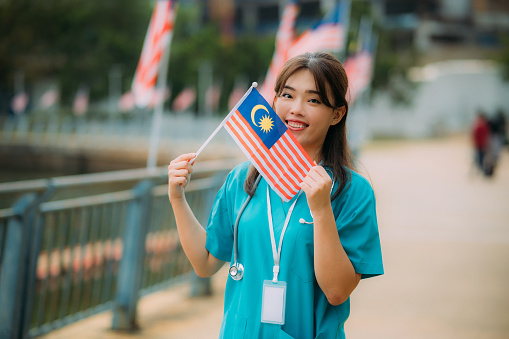 Portrait of a young Asian female nurse holding the Malaysian flag at a public park, celebrating Independence Day and National Day.