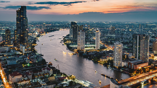 Illuminated glowing Bangkok Cityscape in colorful sunset twilight. High angle long exposure panorama view along the Chao Phraya River into the setting sun at dusk. Hasselblad X2D 102 MPixel Crop. Bangkok, Thailand, Southeast Asia