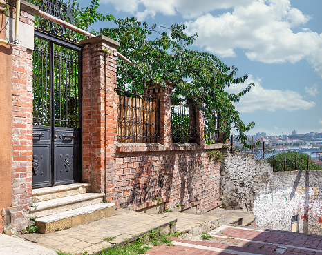 Exterior of old abandoned red bricks fence, wrought iron door and green trees on sunny summer day, Balat district, Istanbul, Turkey