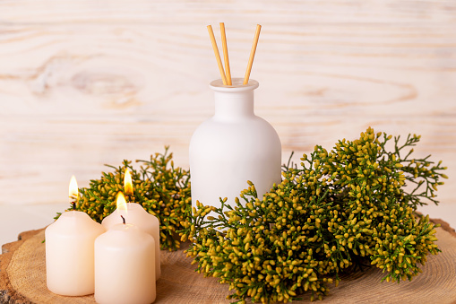 White bottle of aromatic reed diffuser with sticks on wooden background close-up. Burning candles and juniper branches create an atmosphere of calm and relaxation. Spa, rest, recovery