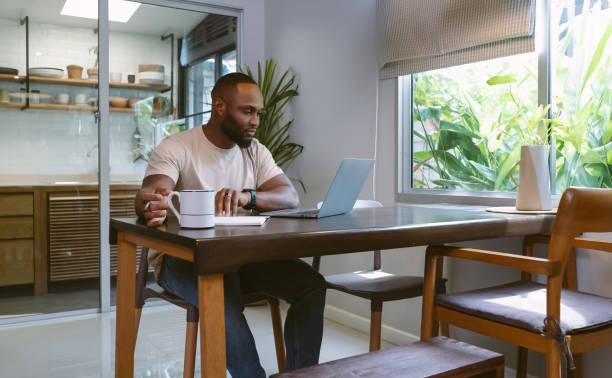 Confident African American bearded man working with laptop computer remotely while sitting at wooden table in kitchen. Black millennial guy do freelance telework at home office