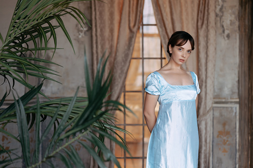 A young beautiful girl in a blue Regency dress stands at a wooden window in the estate. The classic artistic image of Russian literature of the 19th century, outdated.