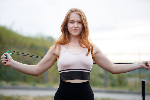 the girl spread her arms in different directions holding a skipping rope outdoors on a summer evening. athletic girl with red hair training