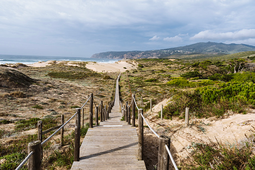 Empty, wooden boardwalk on a beach Praia do Guincho in Sintra. View of grass and sand with hills and Atlantic Ocean in the background.