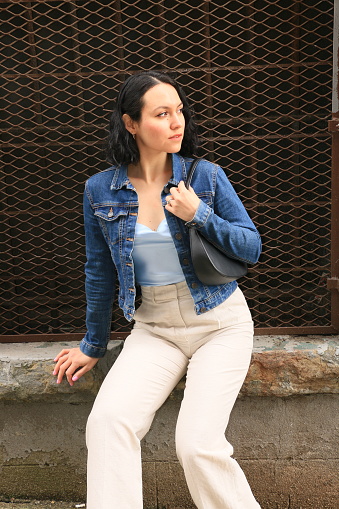 A Mexican model sitting on a stone sill in front of dark security mesh. She is wearing short brown hair, earring, makeup, a blue top, denim jacket and white pants.