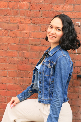 A side view of a happy smiling Mexican woman. She is wearing medium,  length black curly hair, an open blue denim jacket, a blue top and white pants.