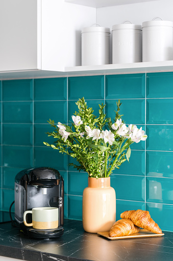 coffee maker with cup, fresh croissants on wooden plate and ceramic vase with blossom flowers on marble countertop at kitchen with cozy interior and blue tile on wall
