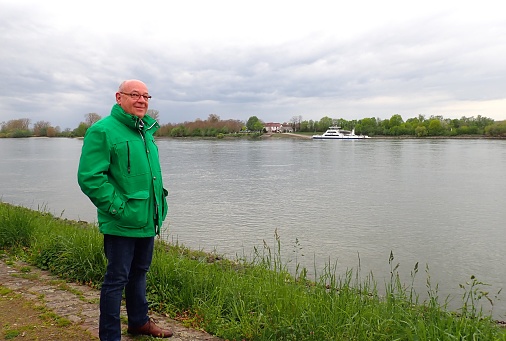 A middle-aged man in a green jacket looks thoughtfully from the shore at the Rhein River, cool cloudy weather.