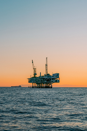 A stunning image of an offshore oil rig at sunset off the coast of Huntington Beach, California. The setting sun highlights the industrial machinery and equipment used in the drilling and extraction of fossil fuels, including crude oil and natural gas. \n\nThis photograph captures the intersection of the energy industry and the natural beauty of the Pacific Ocean, and speaks to issues of fuel and power generation, energy crises, and environmental concerns surrounding the oil and gas industry.