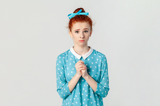 Portrait of hopeful beautiful ginger woman with bun hairstyle, standing with hands together, pleading, asking to forgive, wearing blue dress. Indoor studio shot isolated on gray background.