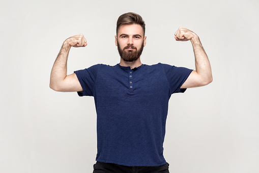 Portrait of strong handsome man with beard wearing dark blue T-shirt looking assertive and raising arms to show biceps, feeling powerful. Indoor shot isolated on gray background.