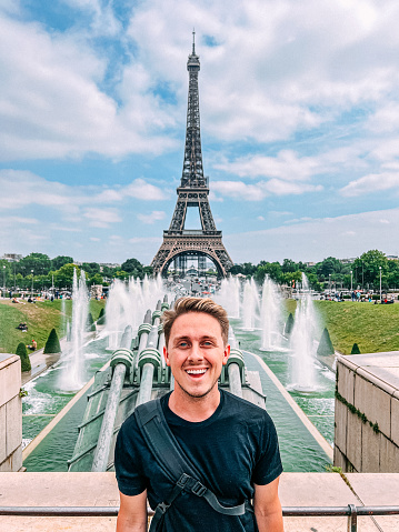 Young Millennial-Age or Gen-Z Handsome Backpacking American Tourist Man Standing at the Trocadero Fountains for a Portrait With the Eiffel Tower in the Background in Paris, France to Post on Social Media. He is wearing a side sling backpack and carrying a local newspaper.