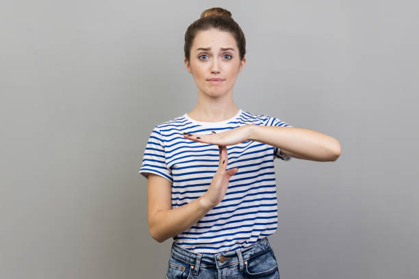 Woman showing time out gesture, looking with imploring eyes, hurry to meet deadline. I need more time. Portrait of woman wearing striped T-shirt showing time out gesture, looking with imploring eyes, hurry to meet deadline. Indoor studio shot isolated on gray background. time out signal stock pictures, royalty-free photos & images