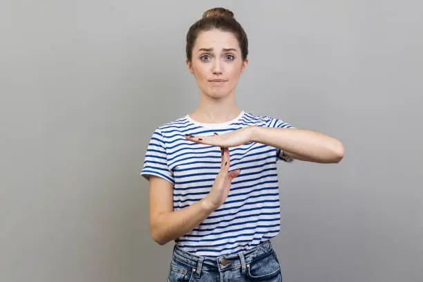 I need more time. Portrait of woman wearing striped T-shirt showing time out gesture, looking with imploring eyes, hurry to meet deadline. Indoor studio shot isolated on gray background.