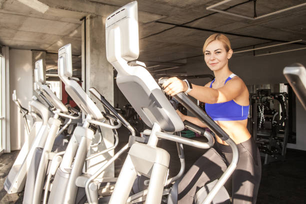donna atletica che usa l'ellittica in un centro fitness, guardando la telecamera e sorridendo. - spinning health club instructor gym foto e immagini stock