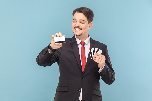 Portrait of delighted smiling greedy man with mustache standing holding many credit cards with big sum of money, wearing black suit with red tie. Indoor studio shot isolated on light blue background.