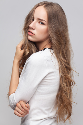 Closeup portrait of adorable attractive young adult woman with long wavy blond hair looking at camera with thoughtful facial expression. Indoor studio shot isolated on gray background.