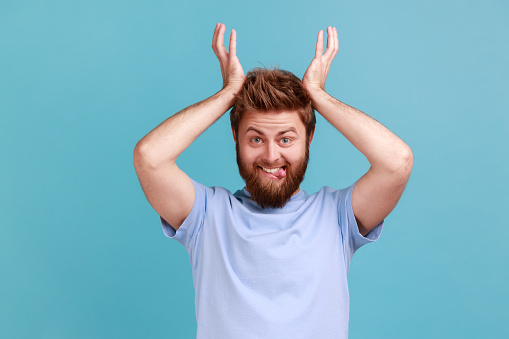 Portrait of positive bearded man standing with antler horns over head and showing tongue out, looking with comical humorous expression. Indoor studio shot isolated on blue background.