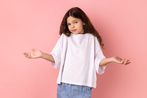 Portrait of confused little girl in white T-shirt shrugging shoulders with no idea gesture, clueless embarrassed face, don't know the correct answer. Indoor studio shot isolated on pink background.