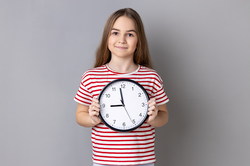 Portrait of smiling little girl wearing striped T-shirt holding big wall clock, looking at camera with pleasant emotions, time to go. Indoor studio shot isolated on gray background.