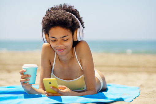 Content young ethnic female traveler with dark Afro hair, in stylish bikini and headphones, drinking takeaway coffee and messaging on smartphone while listening to music and sunbathing on sandy beach