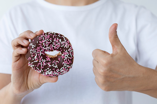 Caucasian woman is showing doughnut with sprinkles  to camera and is doing thumbs up gesture