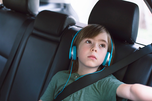 Sad little child with blue headphones listening music while traveling by car. Everyday car journeys.