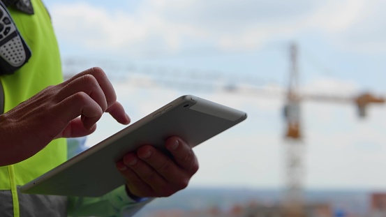 Close up of male hands holding tablet tapping and scrolling on screen outdoors. Man engineer builder in uniform browsing online on device. Male fingers typing on gadget display, technology concept