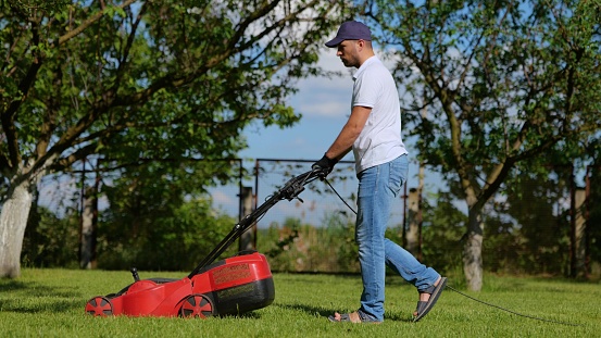 Side view of handsome male worker working in private garden mowing lawn with roller machine. Man walks in the yard and mows green grass with lawn-mower. Gardener job, garden care. Summer day