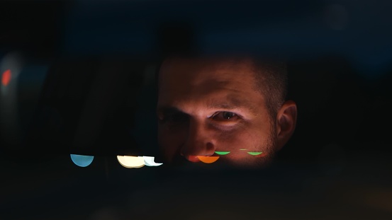 Close up of young serious tired man inside the car in evening. Handsome Caucasian male driving his car and looking at rear view mirror. Blurred background. Night time. Travel. Driver concept