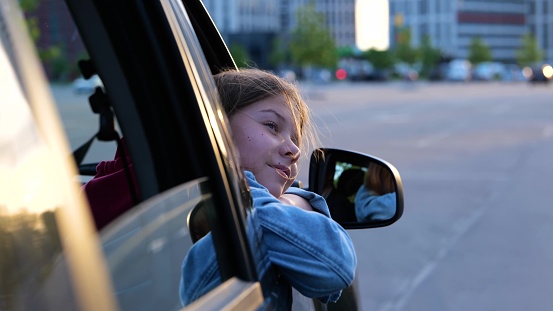 Child in car window. Family trip. Caucasian little girl pulling hand through window. Dreamy teen female looks out of window in vehicle traveling in town. Girl stretches out her hand to wind. Close up